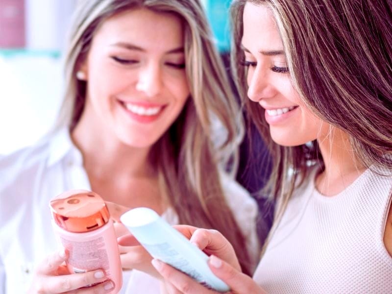 Two women smiling and examining bottles of hair beauty products together, highlighting their interest in quality and ingredients.