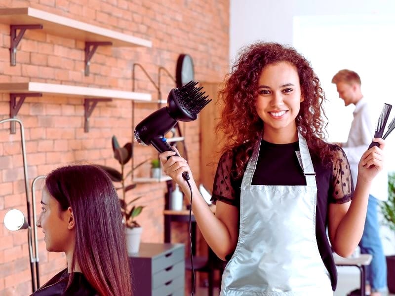 A smiling hairstylist in a modern salon holds a blow dryer and comb, showcasing a clean, professional space focused on non-toxic beauty.