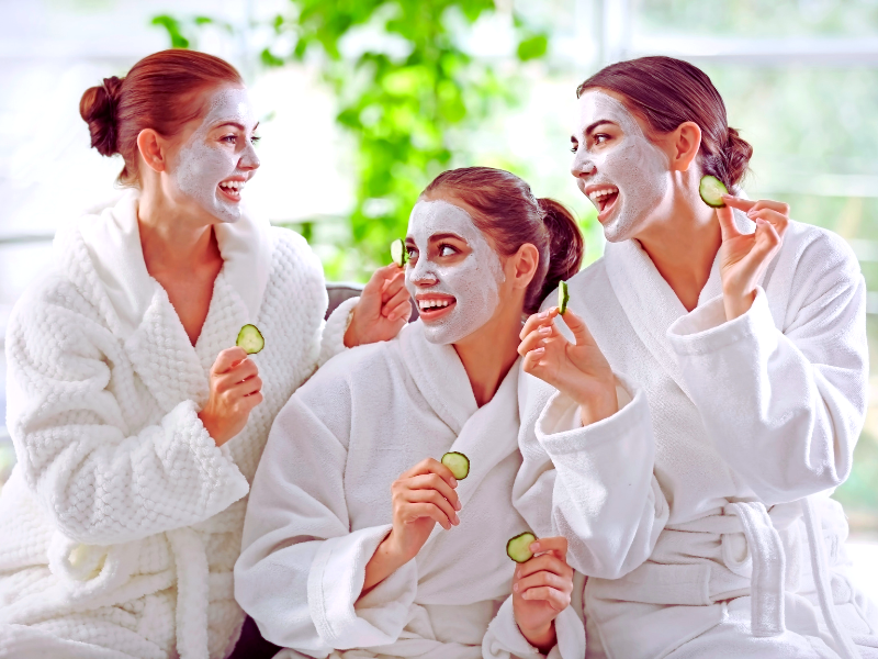 Three women in white robes enjoying a spa day with cucumber slices and face masks, promoting sustainable skincare choices in a natural setting.