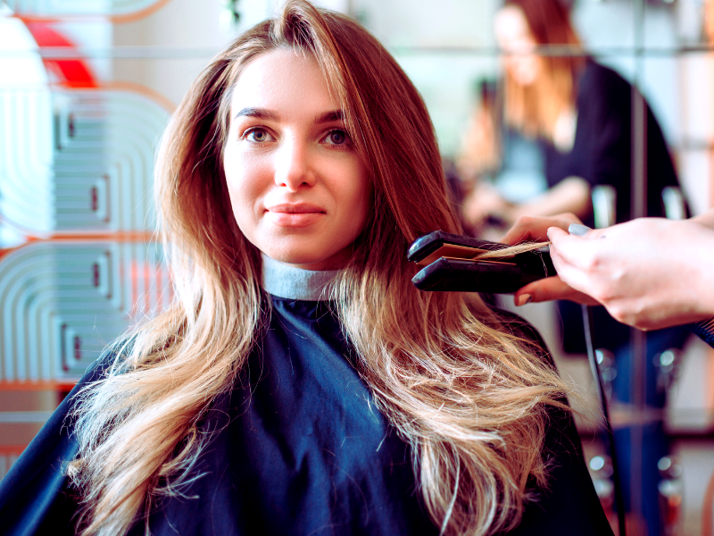 A woman with smooth, healthy hair sits in a salon chair as a stylist uses a flat iron, highlighting professional care and styling.