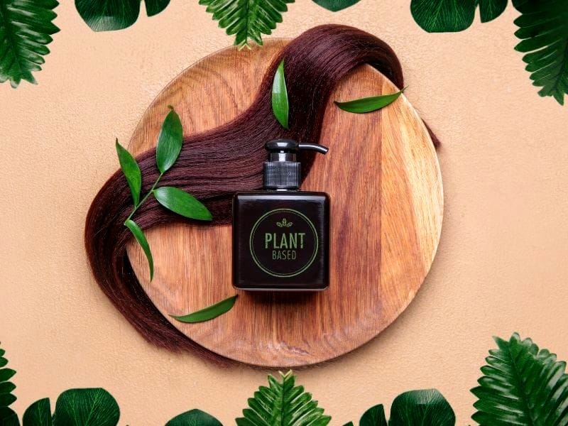 Image featuring a wooden platter with a bottle labeled 'plant-based hair care' surrounded by a strand of shiny brown hair and green leaves.