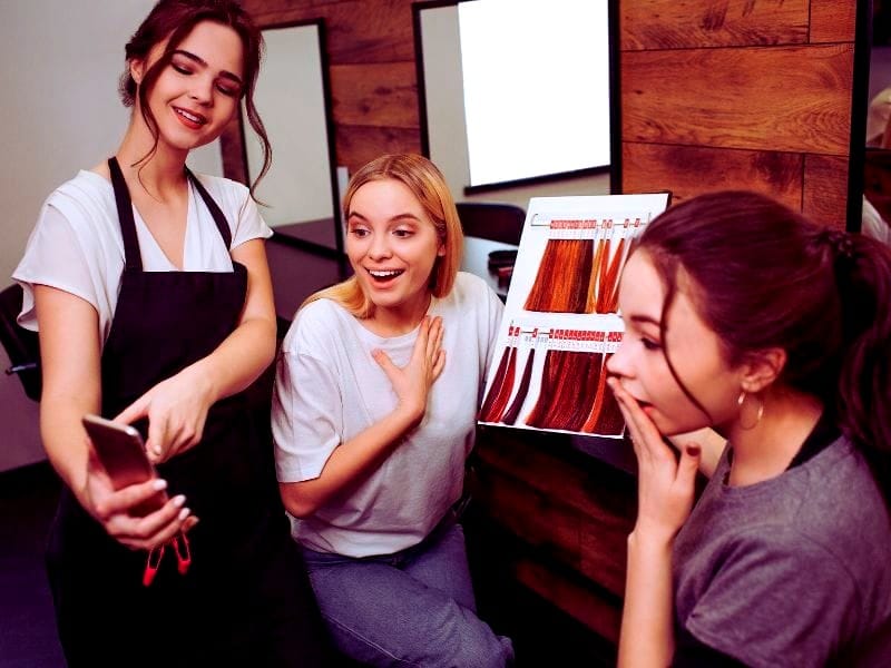 Hair stylist excitedly showing cruelty-free hair products options on her phone to two amazed clients, with a color palette displayed in the salon setting.