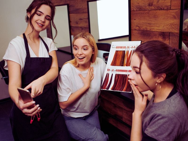 A stylist showing two clients a phone explaining a hair color swatch book in a modern salon, promoting the use of cruelty-free haircare products.