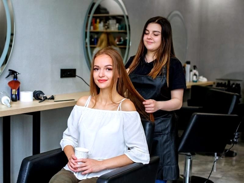 Smiling hairstylist attending to a happy client with long, healthy hair in a modern salon setting.