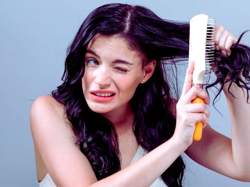 A woman with long, dark, curly hair struggles to brush through tangled strands, making a wincing face while using a wide-tooth comb.