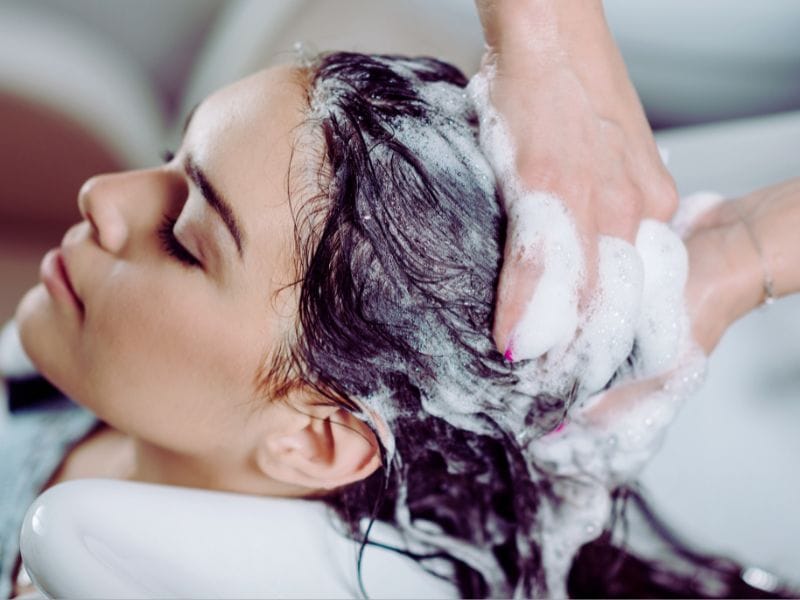 A woman enjoys a relaxing hair wash at a salon, with a stylist massaging shampoo into her scalp, creating a rich, foamy lather.