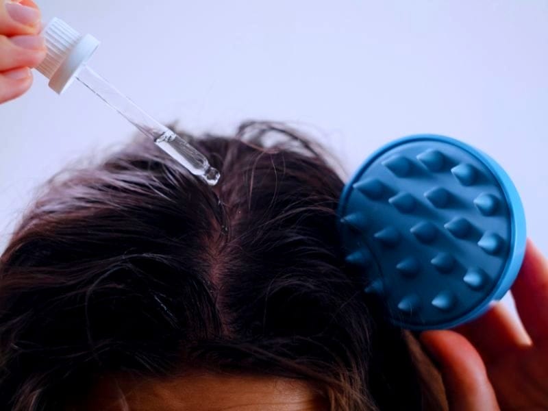 Person applying hair oil with a dropper to the scalp, using a massage brush for better absorption.