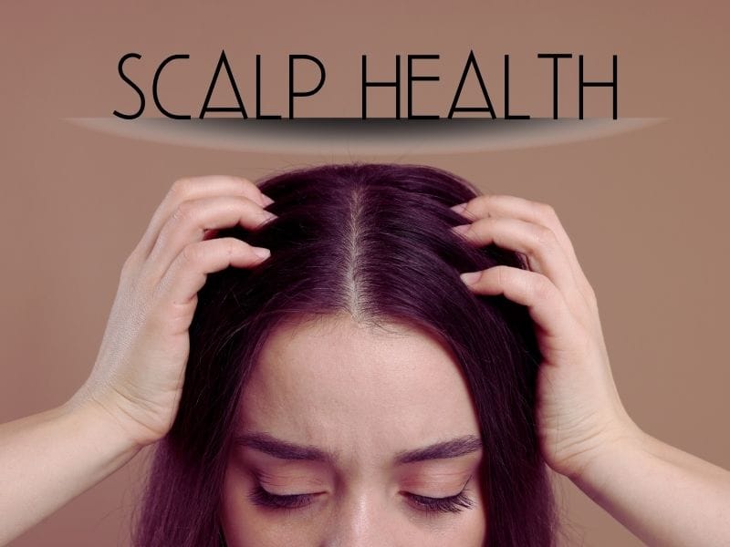 A woman examines her scalp by parting her dark hair with both hands, highlighting scalp health awareness.