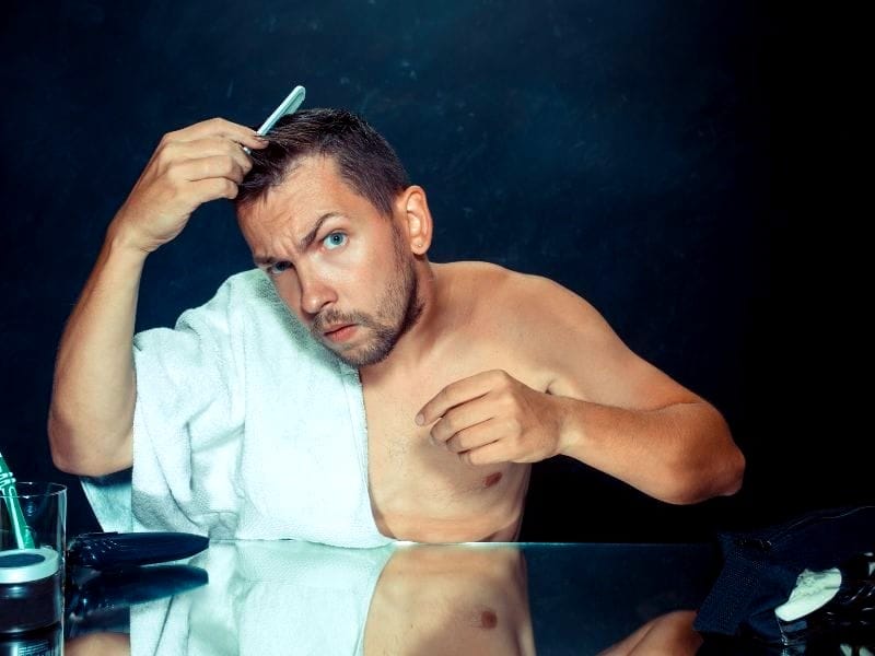 A shirtless man grooming his hair with a comb, focusing on hair care, with grooming essentials on the table.