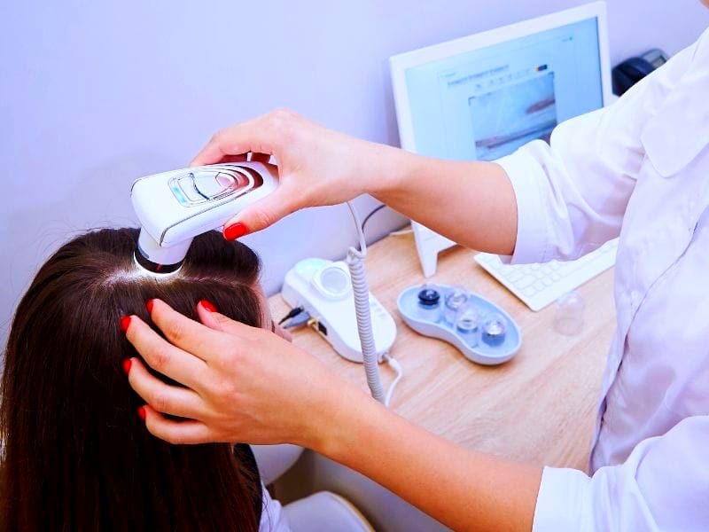 Trichologist examining a woman's scalp with a dermatoscope in a clinic, computer displaying scalp analysis results.