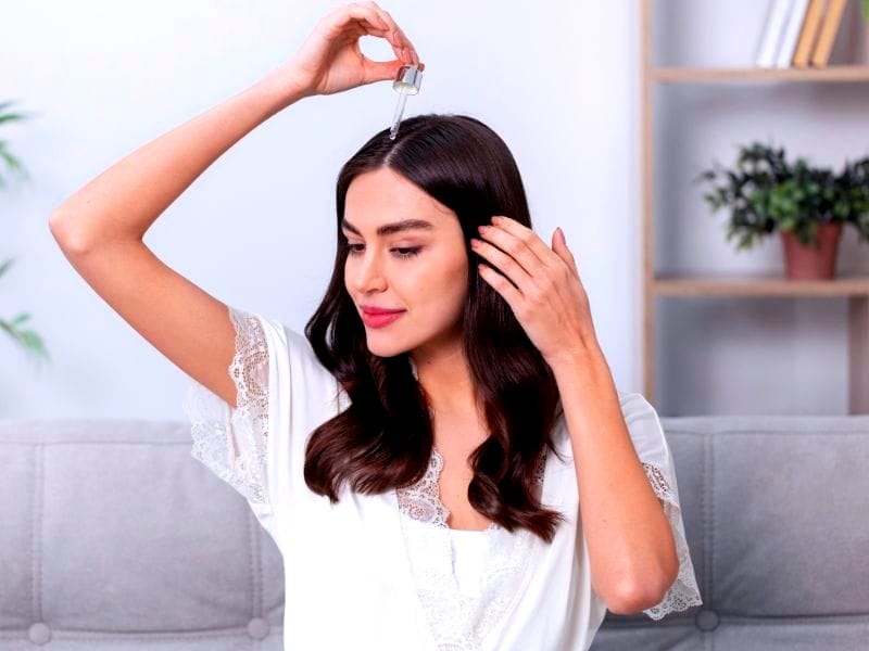 Woman applying hair serum with a dropper on her scalp, sitting on a sofa in a cozy, minimalistic living room.