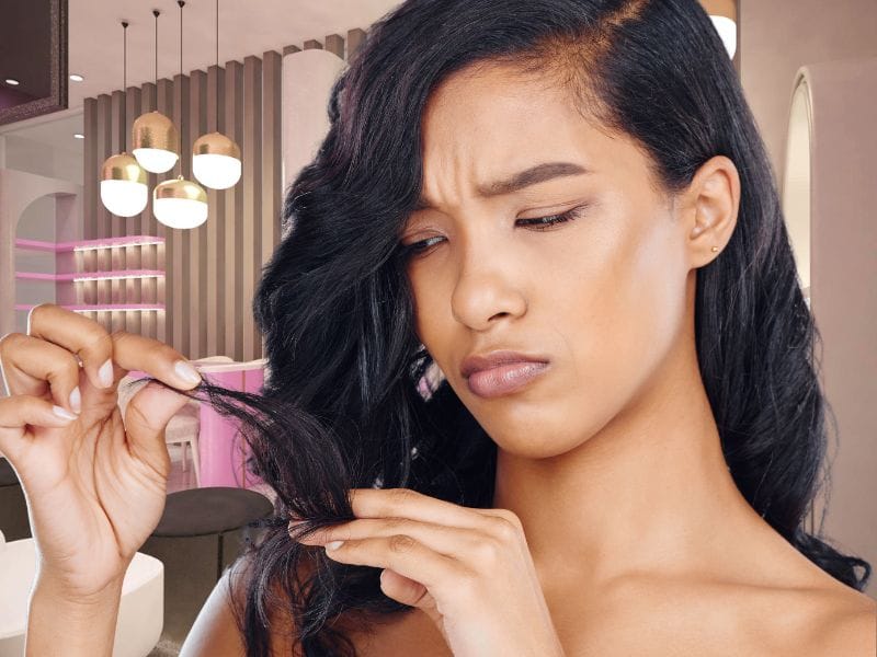 Woman examining dry, damaged hair with a concerned expression in a modern salon setting.