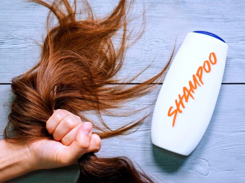 Hand gripping long brown hair next to a white shampoo bottle with orange text "SHAMPOO" – choosing the right formula.