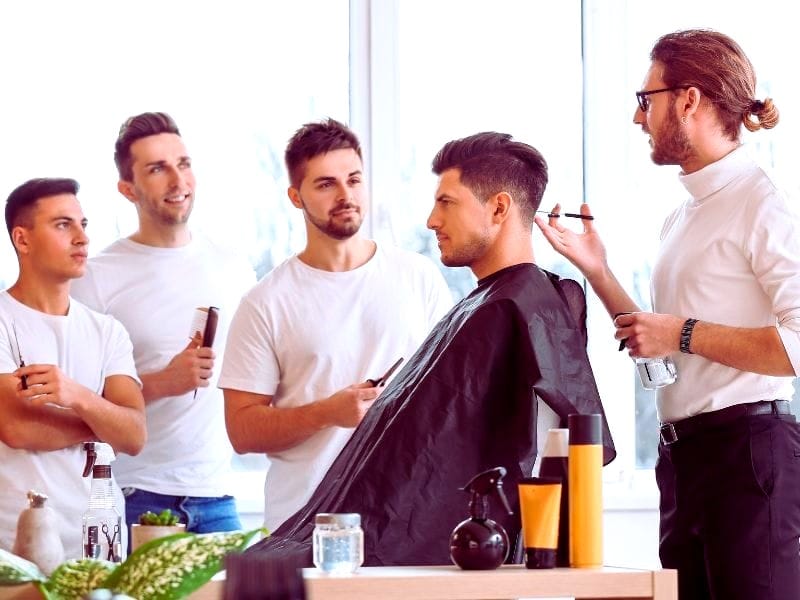 Group of men at a male-friendly salon, one getting a haircut while others observe; professional stylist explaining techniques.