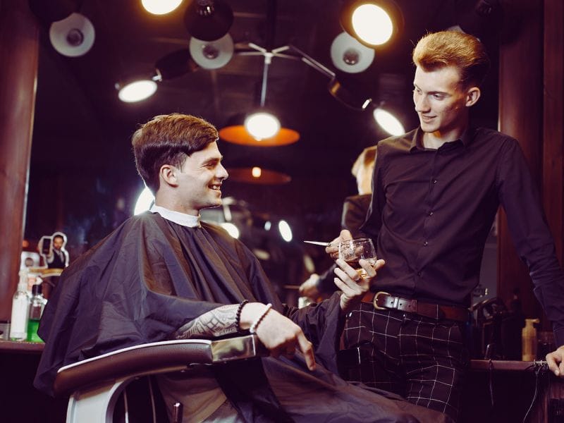 Barber in black attire building trust with a client by offering advice and engaging in friendly conversation in a warm-lit salon.