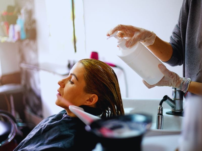 Hairdresser applying a scalp treatment to a client's hair at a salon, using gloves and a white bottle.