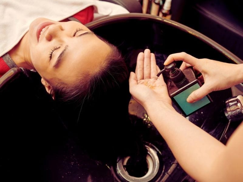 Hair salon scene with a smiling woman receiving a hair treatment; stylist dispensing product into their hand.