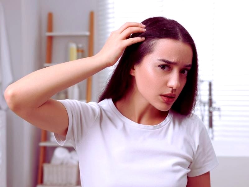 Concerned woman examining scalp and hair for issues like dandruff or oiliness, wearing a white shirt in a bright room.